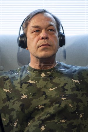 Foto de Un hombre de uniforme militar de 45-50 años, sentado junto a la ventana con auriculares, tratamiento del síndrome postraumático después de la guerra. - Imagen libre de derechos