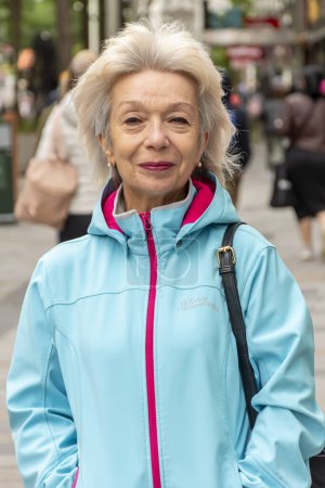 Foto de Retrato de feliz anciana de 60-70 años sobre fondo borroso de la calle, multitud de personas, fondo borroso de la ciudad europea, foto vertical. - Imagen libre de derechos