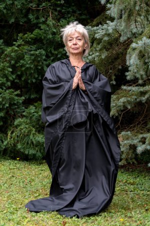 Téléchargez les photos : Une religieuse âgée vêtue d'une robe noire ou d'une soutane sur fond de forêt, les mains pliées en prière. - en image libre de droit