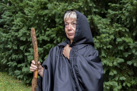 Foto de Una monja anciana en una sotana con un bastón de madera en el fondo del bosque. - Imagen libre de derechos