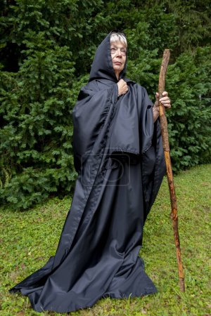 Téléchargez les photos : Femme âgée nonne en soutane noire avec le personnel sur fond de forêt. Pèlerinage et errances. - en image libre de droit
