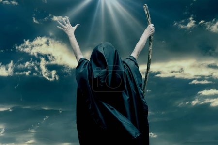 Téléchargez les photos : Une femme vêtue de robes noires leva les mains en l'air, le bâton en bois à la main, le ciel sombre et texturé, illuminé de lumière. Concept : mysticisme et sorcellerie. - en image libre de droit