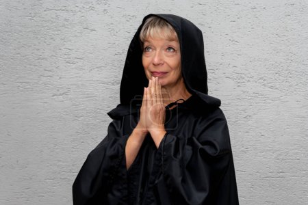 Téléchargez les photos : Portrait d'une religieuse âgée de 60 à 65 ans en soutane noire sur le fond clair du mur. Concept : pèlerinage dans les lieux saints. - en image libre de droit