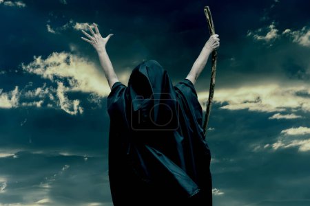 Téléchargez les photos : Femme en robe de moine noir levant les mains, personnel en bois à la main, ciel sombre texturé. Concept : mystique et sorcellerie, prière et concentration, vieille sorcière. - en image libre de droit
