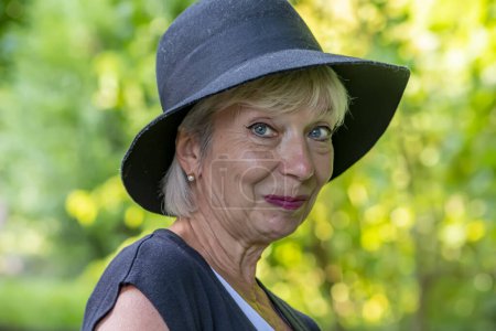 Emotionales Porträt einer älteren blonden Frau mit schwarzem Hut vor verschwommenem Hintergrund der Natur.