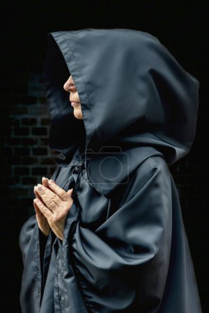 Téléchargez les photos : Vieille religieuse en robe noire contre un mur de briques texturées, mains pliées dans la prière, ton sombre, photo verticale. - en image libre de droit