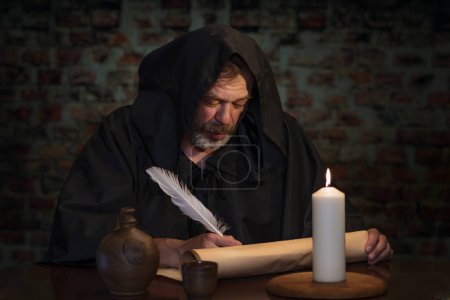 An old monk in his cell writes a message on parchment with a goose quill pen by candlelight.
