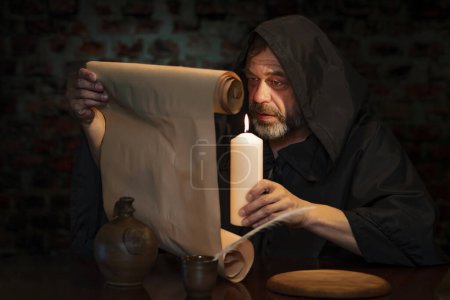 Photo for An old monk reads a message on parchment by candlelight. - Royalty Free Image