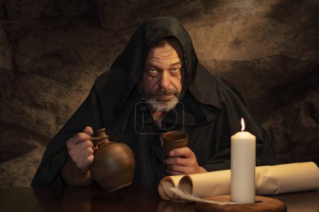 Téléchargez les photos : Portrait émotionnel d'un moine en soutane, gobelet d'argile à la main, assis à une table, une cruche de vin et de boire dans une auberge médiévale. Concept : ivresse et alcoolisme, dîner dans une auberge. - en image libre de droit