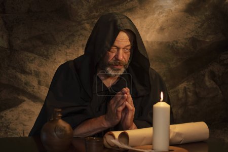 Ein älterer Mönch in schwarzer Soutane betet, die Hände zum Gebet gefaltet, eine dunkle Zelle, eine brennende Kerze auf einem Tisch.