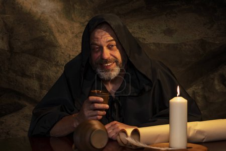 Téléchargez les photos : Joyeux moine en soutane buvant du vin d'un gobelet d'argile assis à une table, une cruche de vin et buvant dans une auberge médiévale. Concept : ivresse et alcoolisme, dîner dans une auberge. - en image libre de droit
