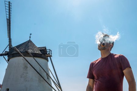 Téléchargez les photos : Un vieil homme fumant une pipe et soufflant de la fumée de tabac sur fond de vieux moulin à vent et de ciel bleu. - en image libre de droit