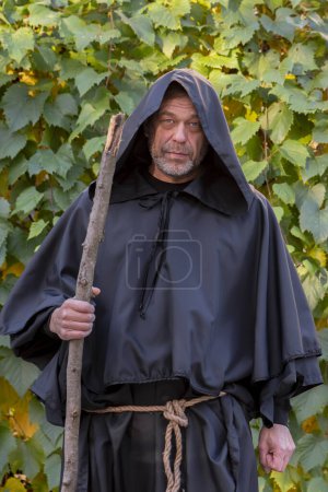 Téléchargez les photos : Portrait d'un moine solitaire avec un bâton en bois sur fond neutre de feuilles de raisin. - en image libre de droit
