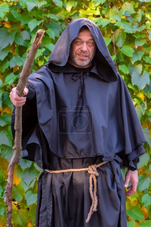 Téléchargez les photos : Moine en robe noire avec un bâton en bois sur le fond des feuilles de raisin. Mysticisme et magie, sorciers et sorcières. production de vin dans un monastère par des moines. - en image libre de droit