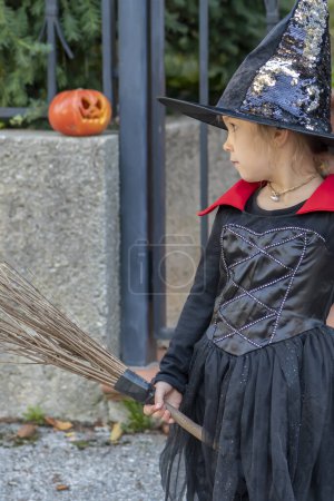 Téléchargez les photos : Fille en costume de sorcière avec balai et chapeau attendant des bonbons à l'entrée de la maison. Halloween de vacances d'automne, citrouille peinte. - en image libre de droit
