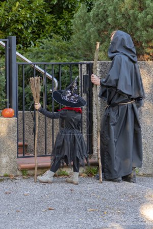 Photo for A girl and a man dressed as a witch and a monk stand at the wicket gate in front of the house waiting for sweets from the owners. Concept: sweets or nasty things, Halloween games and activities. - Royalty Free Image