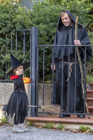 Photo for A man in a monk's robe with a staff giving candy to a girl dressed as a witch, Halloween. - Royalty Free Image
