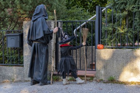 Photo for A girl and her father dressed as a witch and a monk stand at the wicket gate in front of the house waiting for sweets from the owners. Concept: Halloween games and entertainment, festive ritual. - Royalty Free Image