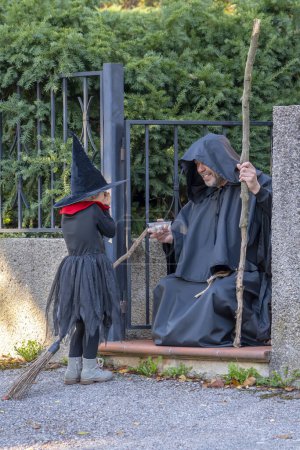 Photo for A girl dressed as a witch with a hat and a broom, a man in a monk's robe with a staff handing out candy, Halloween. - Royalty Free Image