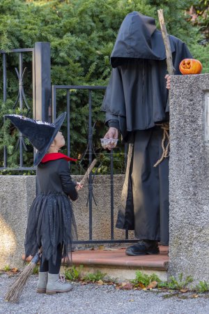 Photo for A man monk treats a girl witch with candy and sweets at the entrance to the house. Autumn Halloween party. - Royalty Free Image