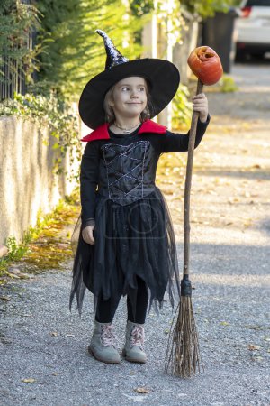 Téléchargez les photos : Une fille avec un balai et une citrouille d'Halloween marche dans la rue en costume de sorcier. - en image libre de droit
