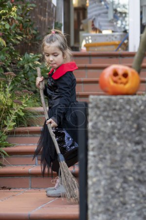 Téléchargez les photos : Une fille habillée en sorcière balaie les poubelles sur les marches avec un balai en bois pour célébrer Halloween. - en image libre de droit