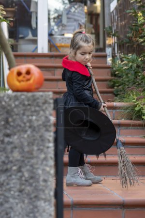 Téléchargez les photos : Portrait d'une petite fille habillée en sorcière avec un balai, balayant un chemin à l'extérieur d'une maison, vacances d'Halloween, citrouille orange. - en image libre de droit