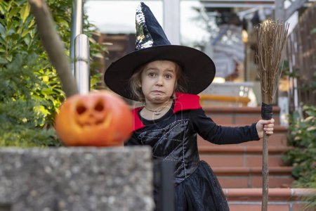 Téléchargez les photos : Portrait émotionnel d'une petite fille blonde en costume de sorcière avec manche à balai, vacances d'Halloween, citrouille orange. - en image libre de droit