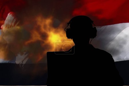 Foto de Un militar con auriculares y portando un portátil contra una bandera yemení, una explosión de fuego y humo. - Imagen libre de derechos