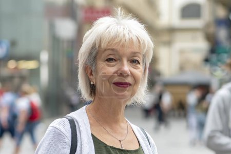 Foto de Una mujer madura de 65-70 años sonríe en un entorno urbano ocupado, mirando a la cámara. - Imagen libre de derechos