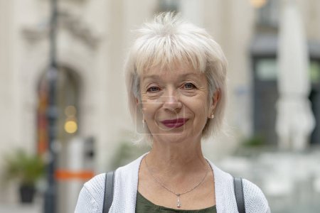 Foto de Retrato callejero de una anciana feliz mujer de 65-70 años sobre un fondo urbano borroso de una calle europea. - Imagen libre de derechos