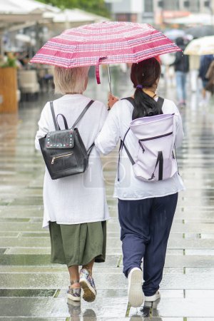 Téléchargez les photos : Une vue arrière de deux femmes sous un parapluie en marchant ensemble sur un trottoir de ville humide sous une pluie légère. - en image libre de droit