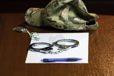 Photo for A military cap, handcuffs and army dog tags, a pen with a white sheet on a wooden table. - Royalty Free Image