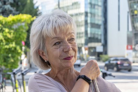 Foto de Una anciana con un reloj de pulsera moderno ajusta una bolsa en su hombro sobre un fondo de edificios modernos. - Imagen libre de derechos