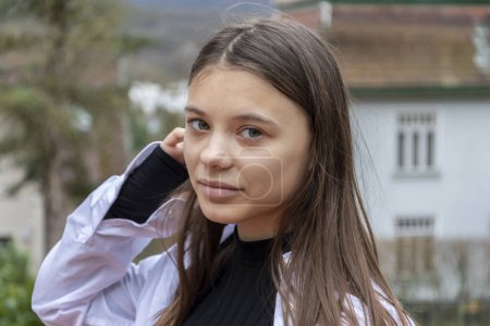 Straßenporträt einer schönen Teenager-Mädchen, 12 Jahre alt, fixiert ihre langen Haare im Hintergrund eines Landhauses, Blick in die Kamera.
