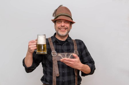 Ein bärtiger Mann mit Tirolerhut deutet auf ein Glas Bier mit Schaum in der Hand auf weißem Hintergrund. Eine Einladung zum Umtrunk.