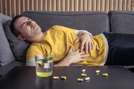Mężczyzna w domowych ciuchach i trzyma ręce na brzuchu leżące na kanapie. stół, na którym znajdują się tabletki, tabletki i szklanka wody. Silny ból brzucha, złe samopoczucie, przeziębienie.