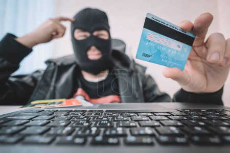 Internetdiebstahl - ein Mann mit Sturmhaube und Kreditkarte, während er hinter einem Laptop sitzt, Geld über das Internet zu stehlen, ist ein Konzept. Kreditkartenbetrug.