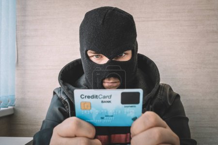 Foto de Delincuente enmascarado calificado usando una tarjeta de crédito robada para comprar cosas en línea. Un ladrón con una máscara negra tiene una tarjeta robada en sus manos. - Imagen libre de derechos