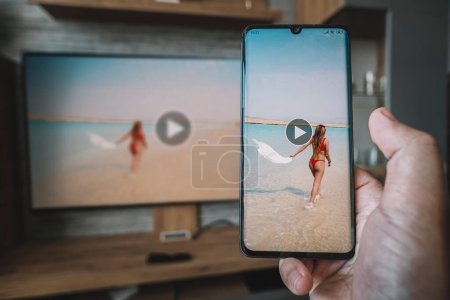 Photo for Male hand holding a smartphone against view of living room with a large screen TV. Use wi fi to transfer images from phone on the monitor screen. Viewing video from your smartphone on your TV. - Royalty Free Image