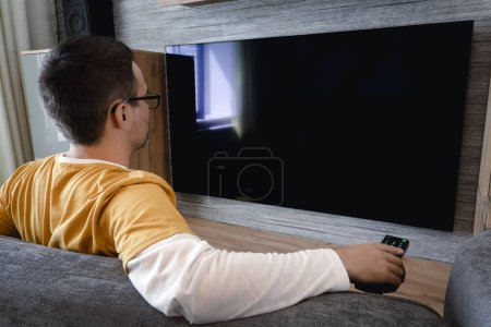 Photo for Man watching television, turning on plasma flatscreen TV-set, pointing remote control at empty TV screen on white wall. Guy switching channels at home, back view. Mockup - Royalty Free Image