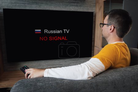 Photo for The concept of disconnecting Russian channels in Europe and the USA. Blocking of Russian TV broadcasting. Young handsome man watching TV on a sofa at home. - Royalty Free Image