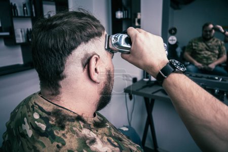 Photo for A young man in a military uniform shaves his head bald for military service. A guy with a beard gets a haircut at a barber shop. - Royalty Free Image