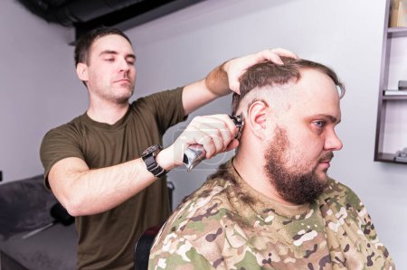 Photo for A young man in a military uniform shaves his head bald for military service. A guy with a beard gets a haircut at a barber shop. shave your bald head - Royalty Free Image