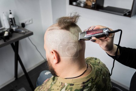 Photo for A young man in a military uniform shaves his head bald for military service. A guy with a beard gets a haircut at a barber shop. - Royalty Free Image