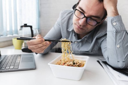 Photo for Young dissatisfied man eating instant noodles while working with laptop in office. Lunch at the office. tasteless junk food - Royalty Free Image