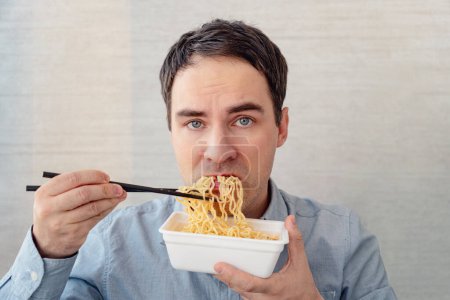 Photo for A young man in a blue shirt is eating noodles from a box with a dissatisfied face. Lunch at the office. tasteless junk food - Royalty Free Image