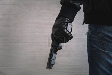 Photo for The concept of the crime of banditry. A dangerous shooter and a black pistol on a dark background. The hired killer is preparing to shoot. Pulls a firearm out of his jacket pocket - Royalty Free Image