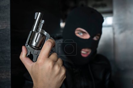 Téléchargez les photos : Un criminel dans une cagoule tient un revolver à canon court dans ses mains et sourit. Concentre-toi sur l'arme. Le concept de crime et de vol. Criminalité de rue. - en image libre de droit