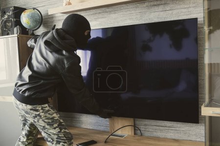 Foto de Un ladrón enmascarado quita un televisor caro de la pared. El concepto de robo de apartamentos. enfoque selectivo - Imagen libre de derechos